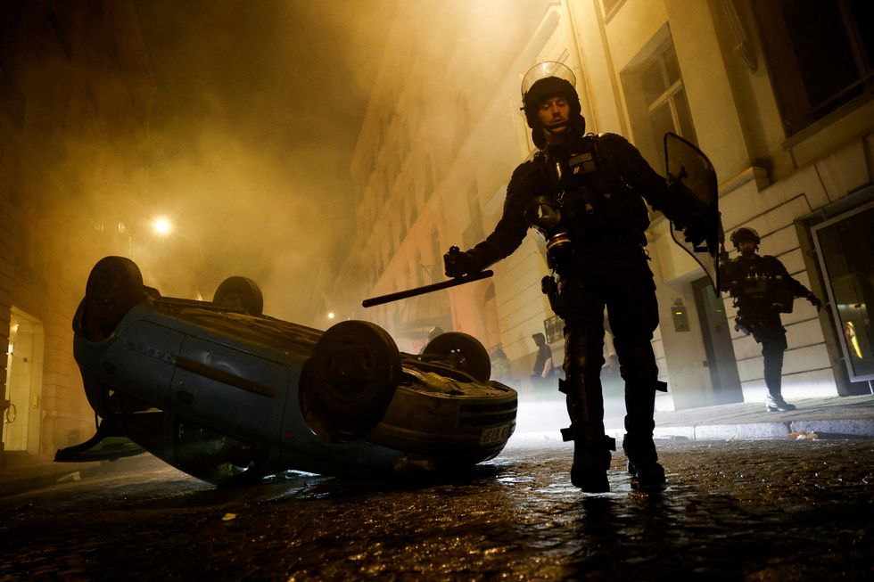 A car turned over with an armed police officer standing next to it