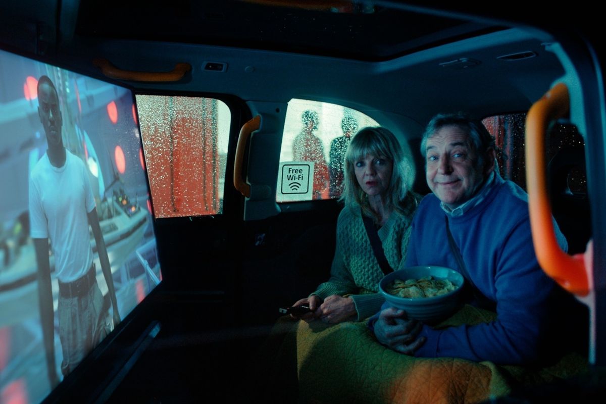a couple sit in the back of a black cab with crisps and a tv remote watching television on freely 