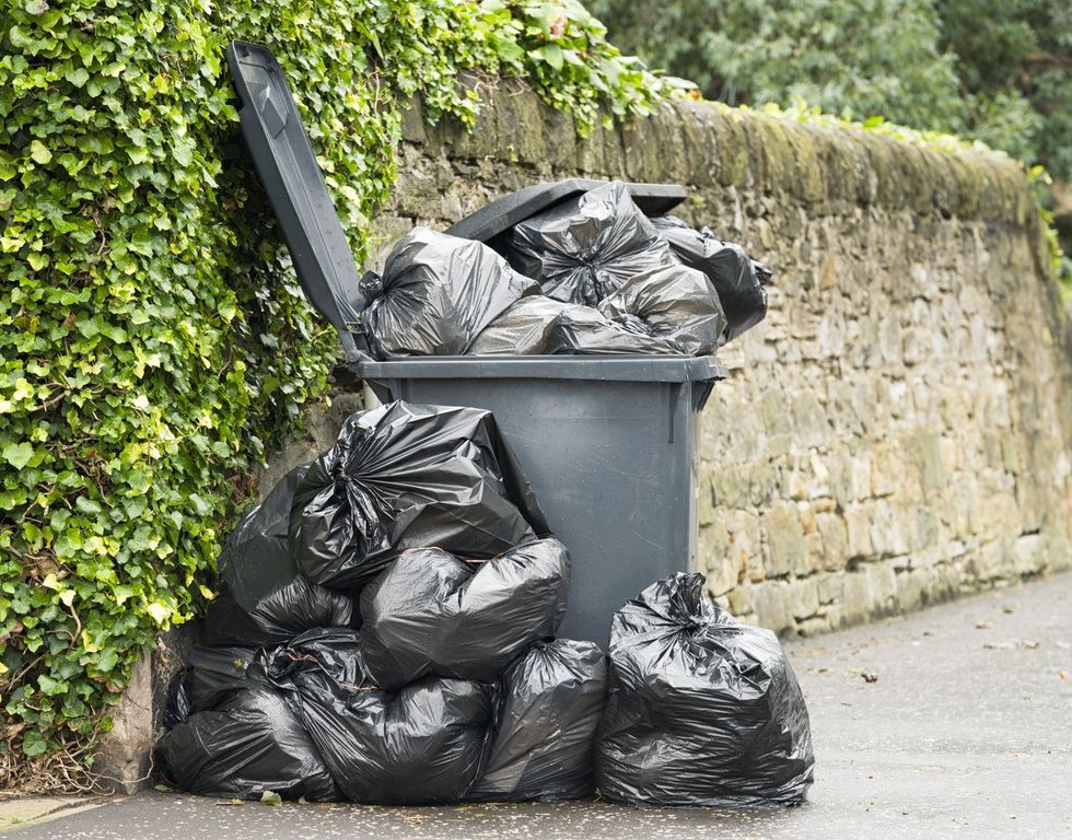 A full wheelie bin with rubbish bags overflowing onto the pavement