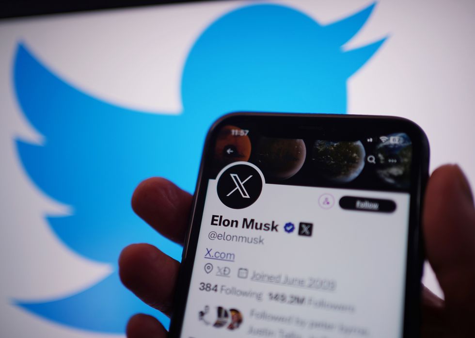 a hand holds a smartphone with elon musks x profile on-screen with the old twitter bird logo in the back ground