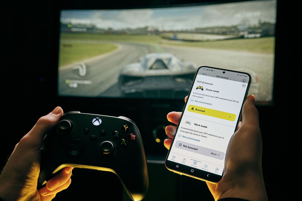 a hand holds an xbox wireless controller with a smartphone enabling game mode with forza in the background from xbox game pass 