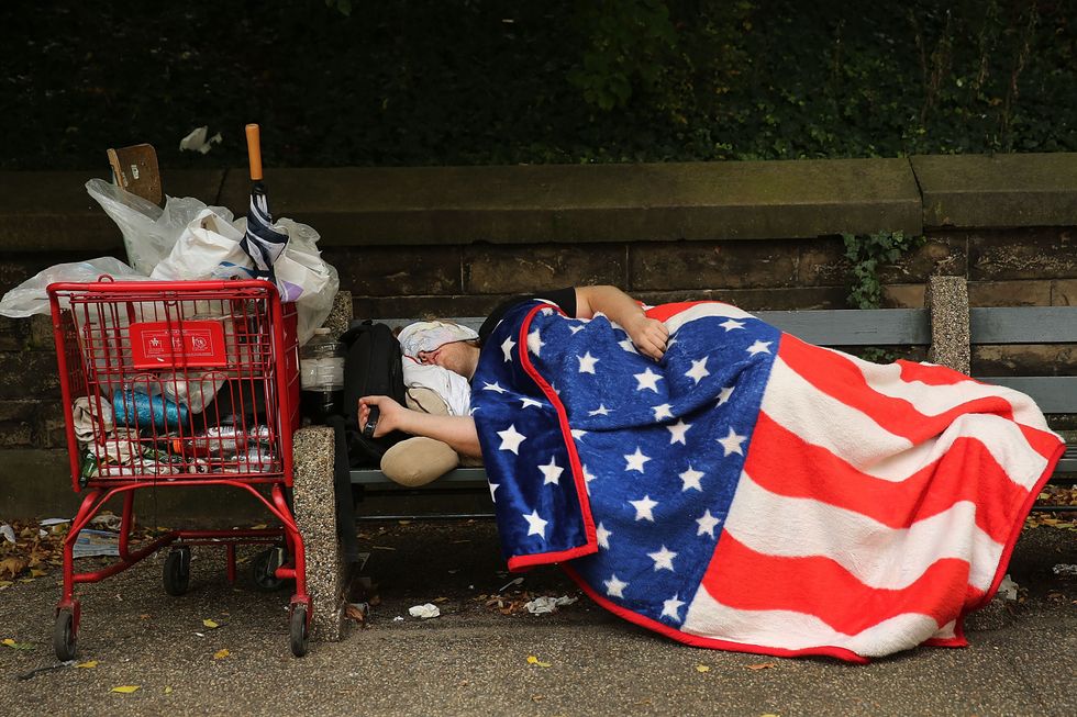 A homeless man sleeps under an American Flag blanket on a park bench