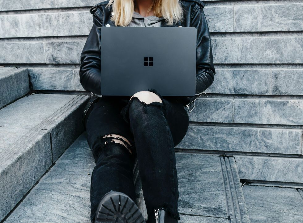 a woman in a leather jacket and jeans sits with a surface laptop on her knees 