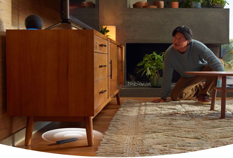 alexa voice remote pro pictured under a entertainment console emitting a ring while a man searches for it