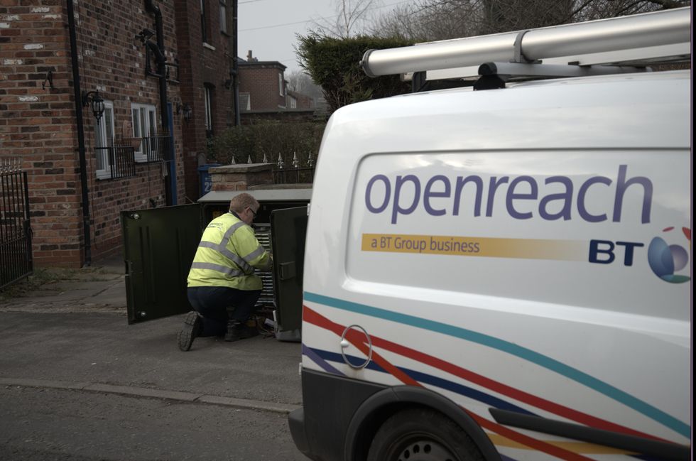 an openreach engineer is pictured making adjustments inside an exchange cabinet on the streets with a BT and openreach branded van parked beside him 