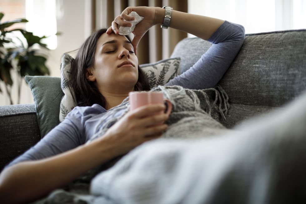 Image of woman with the flu holding her head and wrapped in a blanket on the sofa