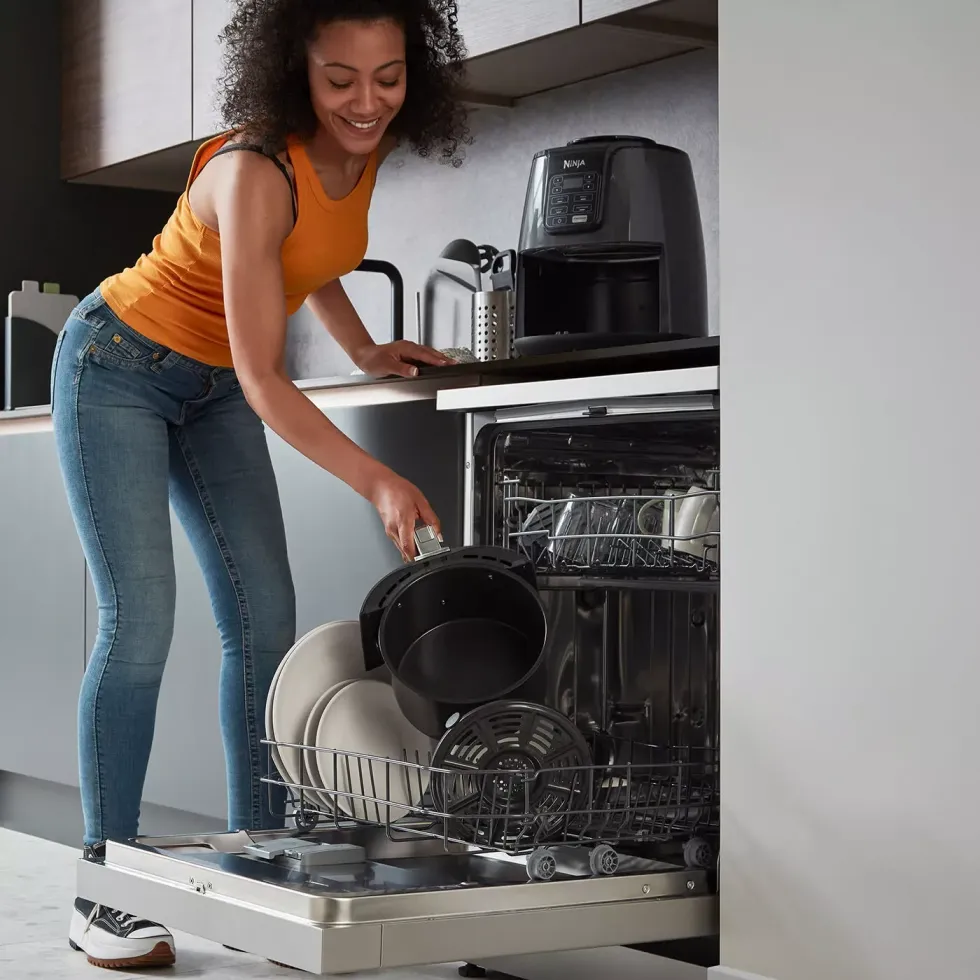 ninja air fryer is pictured being placed in the dishwasher by a woman in jeans and an orange top