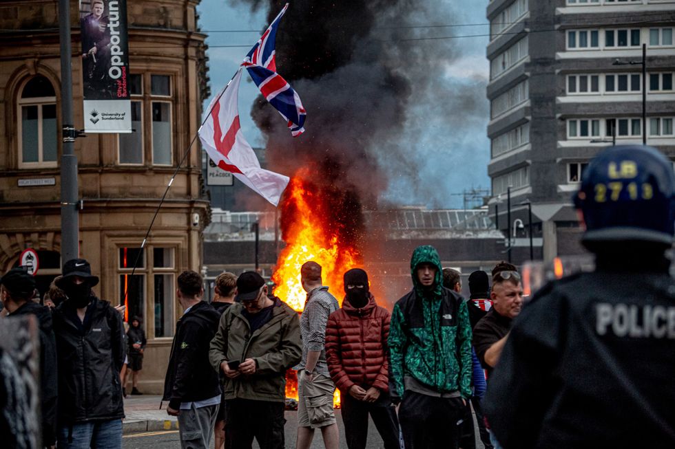 young men stand with their hoods up in front of a burning car in the uk riots